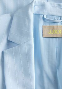 Ανοιχτό γαλάζιο σακάκι με ρίγες και γιακά με σχισμή, που δείχνει εσωτερική μπεζ ετικέτα με κίτρινο κείμενο "JJXX".