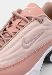 Sneakers Nike Air rose clair avec un tissu texturé, des accents lisses et un logo swoosh argenté bien en vue sur le côté. Lacets ronds inclus.