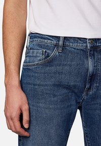 Vasen käsi lepää kyljellä, päällä siniset farkut ja valkoinen t-paita, joka on laitettu sisään.