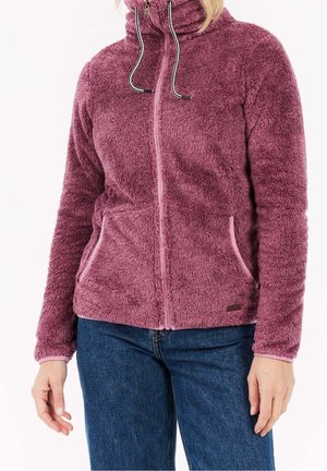 Personne portant une veste polaire mauve, duveteuse, zippée avec des cordons, associée à un jean bleu, debout devant un fond uni.