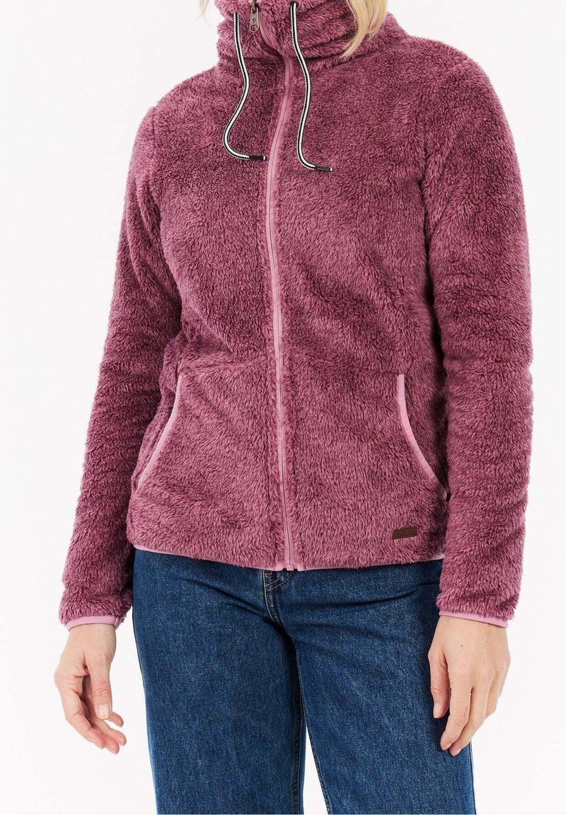 Personne portant une veste polaire mauve, duveteuse, zippée avec des cordons, associée à un jean bleu, debout devant un fond uni.