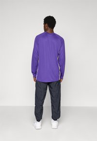 Mand iført en lilla langærmet skjorte med gule detaljer, sorte bukser og hvide sneakers, stående med ryggen til mod en ensfarvet baggrund.
