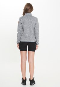 Veste zippée grise en tissu texturé, dotée d'un col haut et d'un patch logo sur l'épaule, accompagnée de shorts noirs et de chaussures.