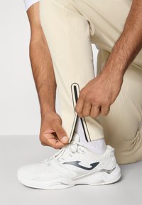 Pantalons de sport de couleur crème avec un détail zippé au niveau du poignet, associés à des baskets blanches. Les baskets ont un logo bleu et une semelle texturée.
