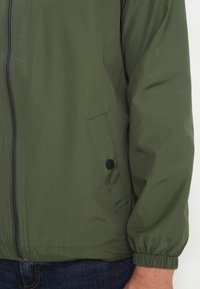 Homme portant une veste zippée verte avec une poche latérale à bouton et des poignets élastiques, associée à un jean bleu.