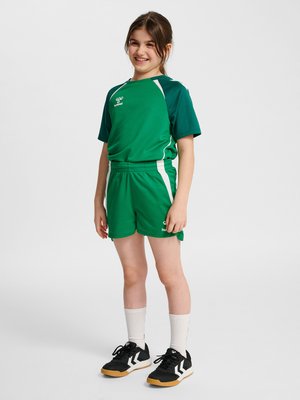 Smilende pige stående iført grønne sports-shorts og T-shirt, hvide sokker og sorte atletiksk sko på ensfarvet hvid baggrund.