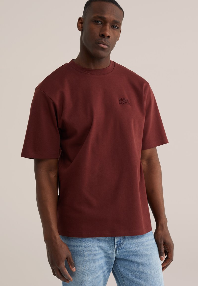 Hombre con camiseta de manga corta color vino con el texto "URBAN BLOOM" en el pecho y jeans azul claro, de pie frente a un fondo liso.