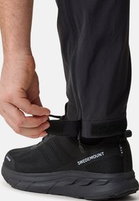 Schwarze Outdoor-Hosen mit Reißverschluss und verstellbarem Klettverschluss am Saum, kombiniert mit schwarzen Sportschuhen, die strukturierte Muster und Markenlogo aufweisen.