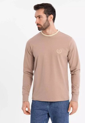 Beige långärmad T-shirt med krämfärgad halsringning, med en liten logotyp på bröstet. Kombinerad med denimjeans.