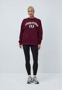 Γυναίκα με μπορντό oversize sweatshirt με το κείμενο "LEGER STUDIO", μαύρα κολάν, λευκές κάλτσες και γκρι αθλητικά παπούτσια, που στέκεται μπροστά σε απλό φόντο.