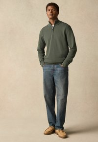 Grüner Pullover mit Viertel-Reißverschluss und strukturiertem Design, kombiniert mit hellblauen Jeans und beigen Schuhen, die Schnürsenkel und eine robuste Sohle haben.
