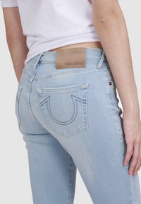 Jean en denim bleu clair avec un design ajusté, arborant une étiquette en cuir avec le logo "True Religion" et une couture distinctive sur les poches arrière.