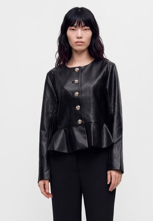 Un mannequin aux longs cheveux foncés porte une veste en cuir noire avec des boutons dorés et un basque, assortie à un pantalon noir.