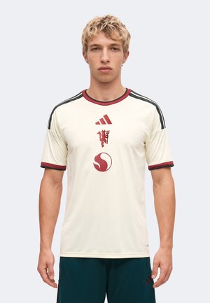 Joven con camiseta de fútbol crema con logos rojos y franjas negras en los hombros, de pie mirando hacia adelante sobre fondo liso.