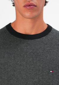Schwarzer und grauer Herringbone-Musterpullover mit rundem Hals, der ein kleines Logo in Rot, Weiß und Blau am unteren Ende aufweist.