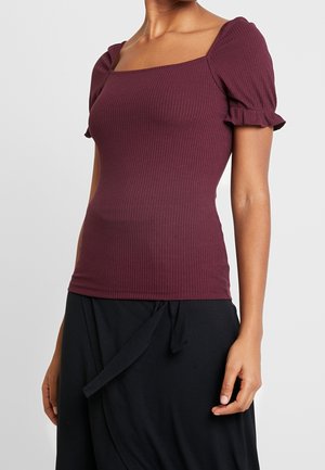 Kobieta w dopasowanym, prążkowanym, fioletowym topie z bufkami na krótkich rękawach i kwadratowym dekoltem, w połączeniu z czarną spódnicą owijaną.