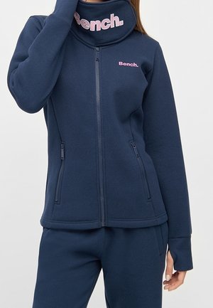 Vrouw draagt een marineblauwe jas met rits en broek met een roze "Bench."-logo op de borst en een nekkraag, hand raakt haar gezicht aan.