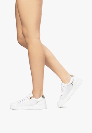 Jambes portant des baskets blanches avec des accents métalliques, un pied à plat sur le sol et l'autre levé comme pour faire un pas en avant.