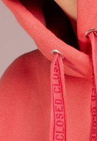 Koraalkleurige hoodie met een zachte textuur, veters met de tekst "CLOSED" bedrukt en zilveren oogjes, met een relaxte pasvorm en capuchonontwerp.