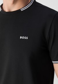 Chemise noire à manches courtes avec col et manches bordés de blanc, avec texte blanc en majuscules "BOSS" sur la poitrine gauche.
