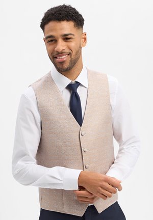 Mann lächelt, trägt eine beige gemusterte Weste, ein weißes Hemd und eine dunkelblaue Krawatte vor einem schlichten weißen Hintergrund.