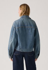 Blauwe denimjas met een ontspannen pasvorm, knopenmanchetten en een kraag. Verweerde details aan de zoom en naden, gecombineerd met donkere jeans.