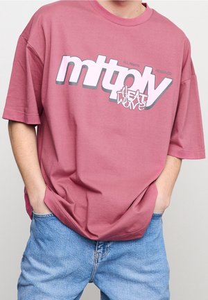 Różowy bawełniany t-shirt z wyrazistym nadrukiem z napisem "multiply HEAT WAVE", z krótkimi rękawami i luźnym krojem. Noszony z niebieskimi dżinsowymi szortami.