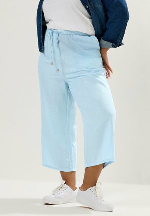 Osoba ubrana w jasnoniebieskie, szerokie spodnie o długości do połowy łydki z troczkiem, białe sneakersy, biały top i dżinsową kurtkę, stojąca z jedną ręką na biodrze.