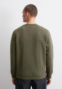 Olívazöld pulóver kerek nyakkal, hosszú ujjal és bordás mandzsettákkal. Az anyag lágy kinézetű, sima textúrával és minimális varrási részletekkel rendelkezik.