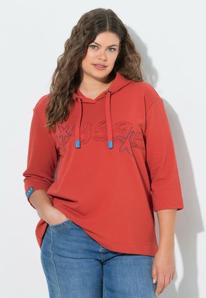 Kvinde med langt bølget hår iført rød hættetrøje med søstjerneudseende og blå jeans, stående med den ene hånd i lommen mod en hvid væg.