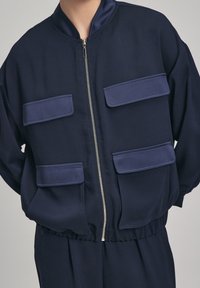 Marint bomberjacka i ett slätt tyg med dragkedja fram, med fyra platta fickor och en ribbad krage. Rak skärning med elastisk midja.