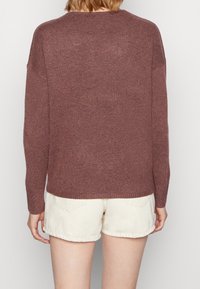 Långärmad, vinröd tröja med lös passform, stickad textur och ribbad nederkant. Bärs över ljusa shorts, vilket visar en enkel design på baksidan.