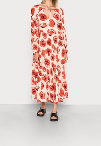 Femme portant une longue robe crème avec de grands motifs floraux rouges, des manches longues, un décolleté en trou de serrure et des sandales à talons ouverts noires.