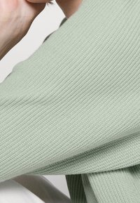 Maglione lavorato a maglia verde menta con texture a coste, vestibilità larga e tessuto morbido. Primo piano che mette in risalto il dettaglio del manico e la texture.