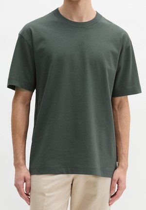 Hombre con camiseta lisa de manga corta de cuello redondo verde oscuro y pantalones beige sobre un fondo blanco.