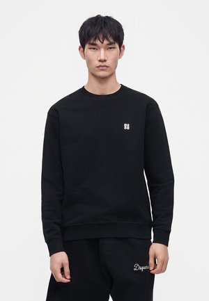 Jeune homme portant un sweat-shirt noir et un pantalon noir avec de petits logos blancs sur la poitrine et la cuisse supérieure, debout devant un fond blanc.