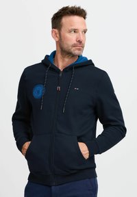 Felpa con zip blu navy, con cappuccio interno blu, caratterizzata da un logo rotondo, coulisse e tasche frontali. Tessuto morbido e vestibilità comoda.