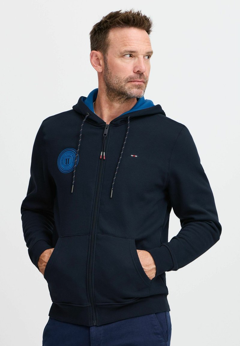 Felpa con zip blu navy, con cappuccio interno blu, caratterizzata da un logo rotondo, coulisse e tasche frontali. Tessuto morbido e vestibilità comoda.