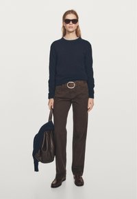 Ciemnoniebieski sweter, brązowe spodnie o szerokich nogawkach i brązowe skórzane buty. Trzyma ciemną torbę z granatowym swetrem zarzuconym na nią. Na nosie okulary przeciwsłoneczne.