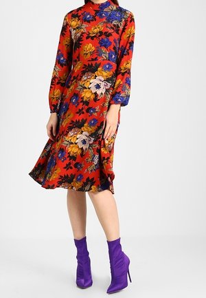 Kvinde iført en rød midikjole med blomsterprint og lange ærmer, kombineret med lilla ankelstøvler med høje hæle, foran en ensfarvet hvid baggrund.