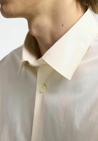 Camicia beige con bottoni e colletto classico, caratterizzata da una texture liscia e una leggera lucentezza. Chiusura con bottoni visibile, con cuciture ben definite.