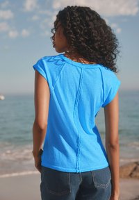Blue short-sleeve T-shirt with a relaxed fit and visible stitching details. Paired with dark denim jeans. Ocean backdrop.