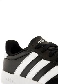 Baskets noires en cuir synthétique avec trois rayures blanches, présentant une languette et des lacets noirs, ainsi qu'une semelle en caoutchouc blanche.