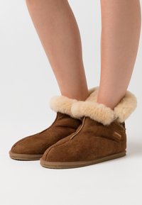 Brown suede ankle boots with fluffy cream-colored wool lining worn on bare legs against a plain white background.