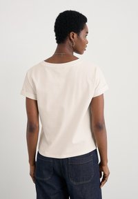 Camicetta corta beige chiaro con una vestibilità rilassata, scollo rotondo, maniche arrotolate e tessuto di cotone morbido. Indossata con pantaloni in denim scuro.
