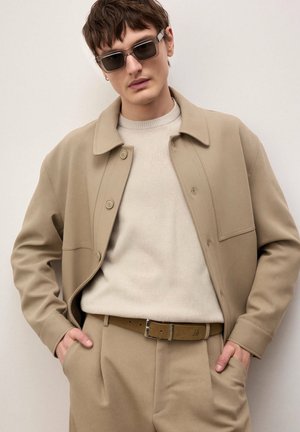 Man wearing beige jacket, matching trousers, light sweater, brown belt, and dark sunglasses, standing with hands in pockets.