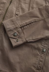 Rjava bombažna jakna z zadrgo spredaj, manšetami na gumbe in kovinskimi poudarki. Ima vidne šive in gladko teksturo.