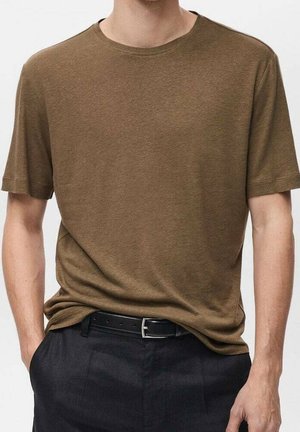 Mand iført en brun t-shirt med korte ærmer og rund hals, let stoppet ind i sorte bukser med sort bælte, hænderne i lommerne.
