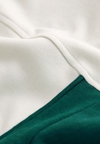 Tissu en coton écru et vert, présentant une texture lisse avec des coutures visibles et un détail en bloc de couleur diagonale.