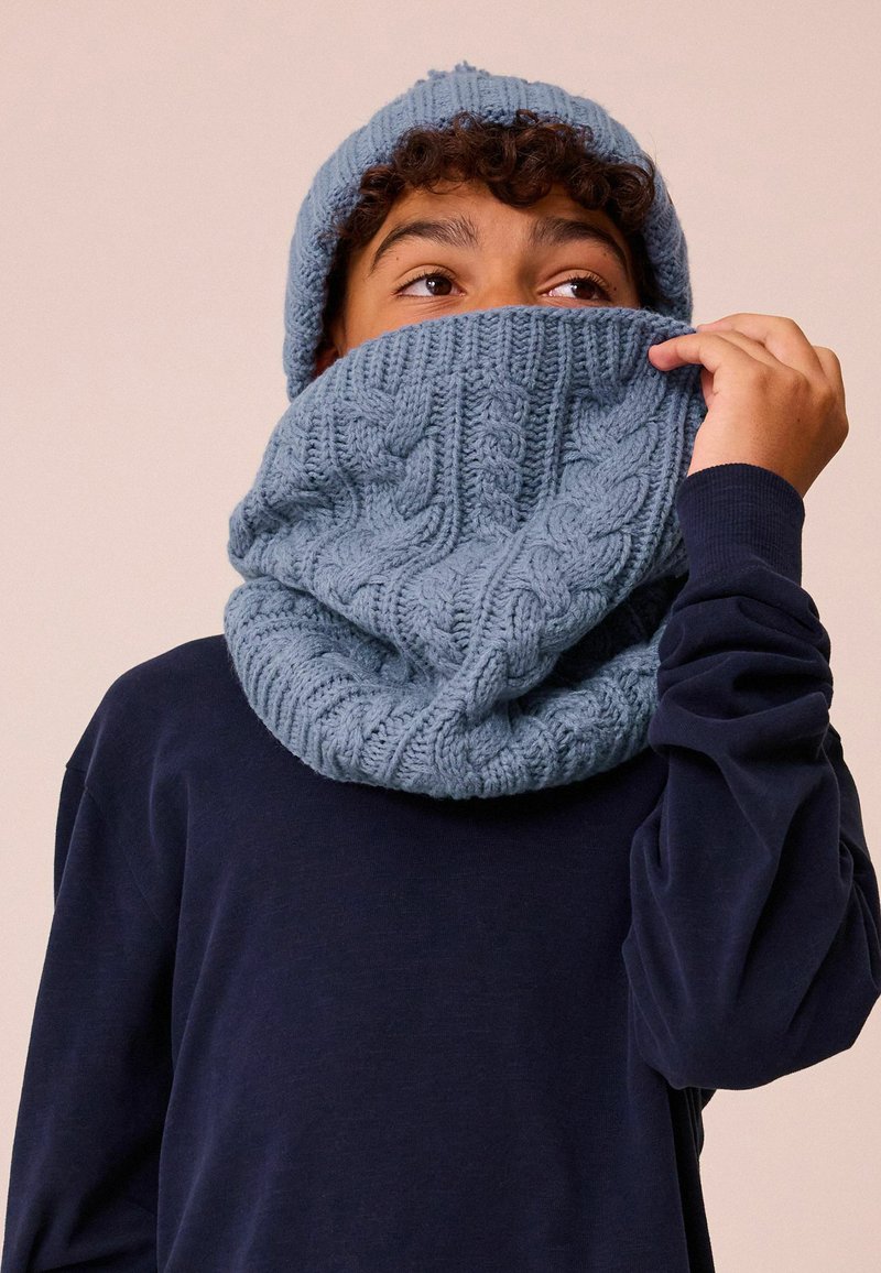 Berretto lavorato a maglia blu e collo abbinato con un motivo a treccia, indossati insieme da una persona in una maglietta scura a maniche lunghe. Texture morbida evidente.